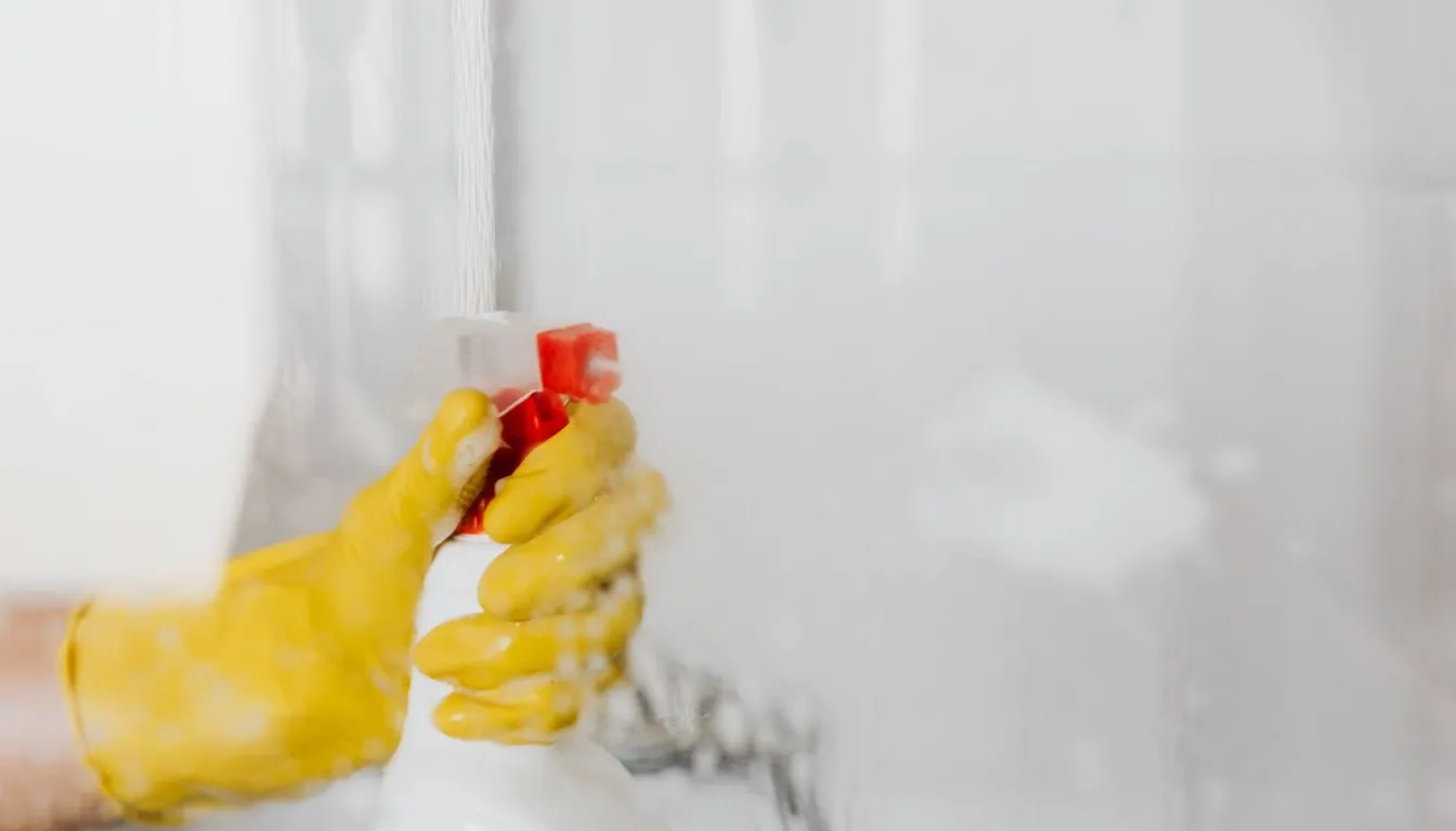 Gloved hand spraying cleaner onto a glass shower wall
