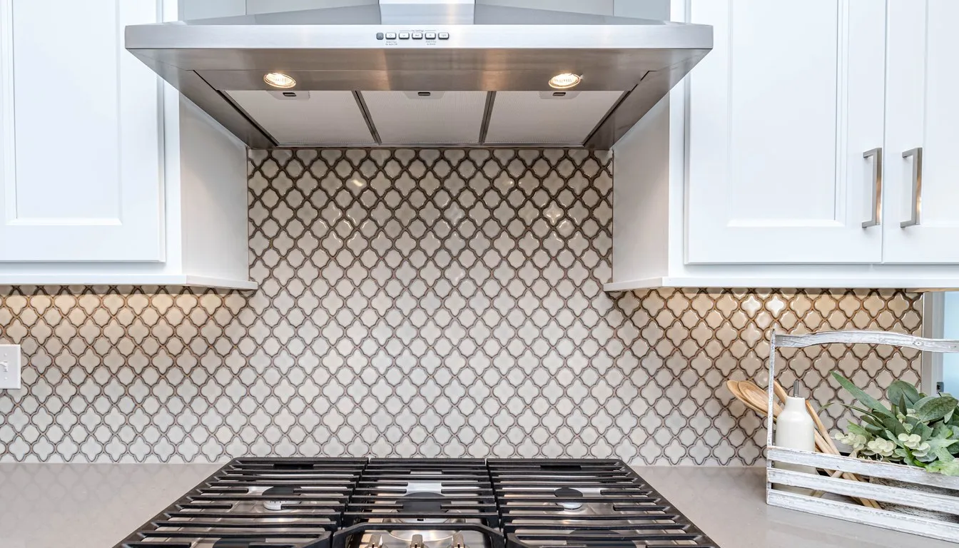 Stainless steel chimney-style range hood with its lights on, mounted above a gas cooktop and a tiled backsplash between white kitchen cabinets